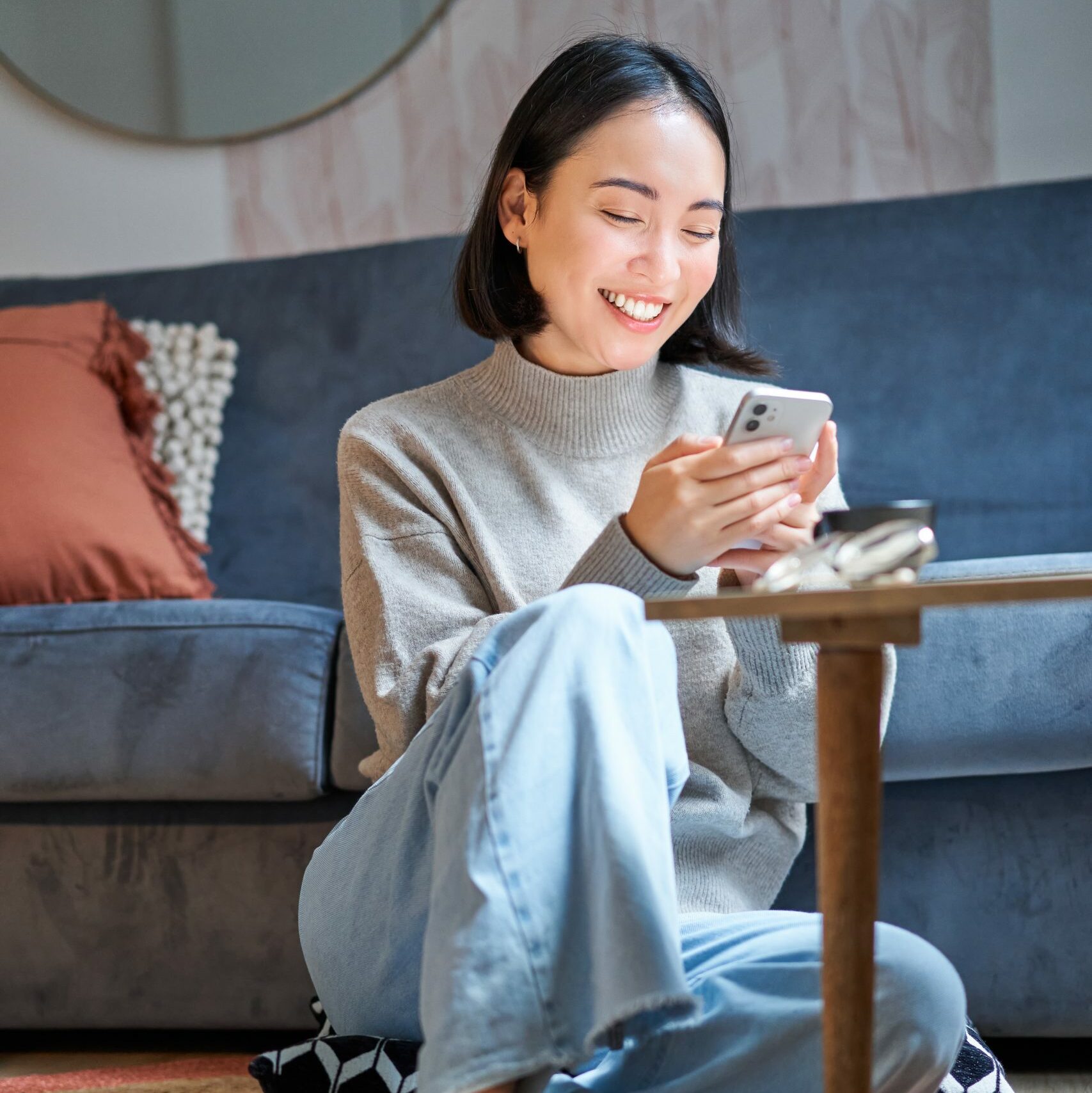 Vertical shot of stylish korean woman sitting on floor at home, using her mobile phone, holding smartphone and smiling